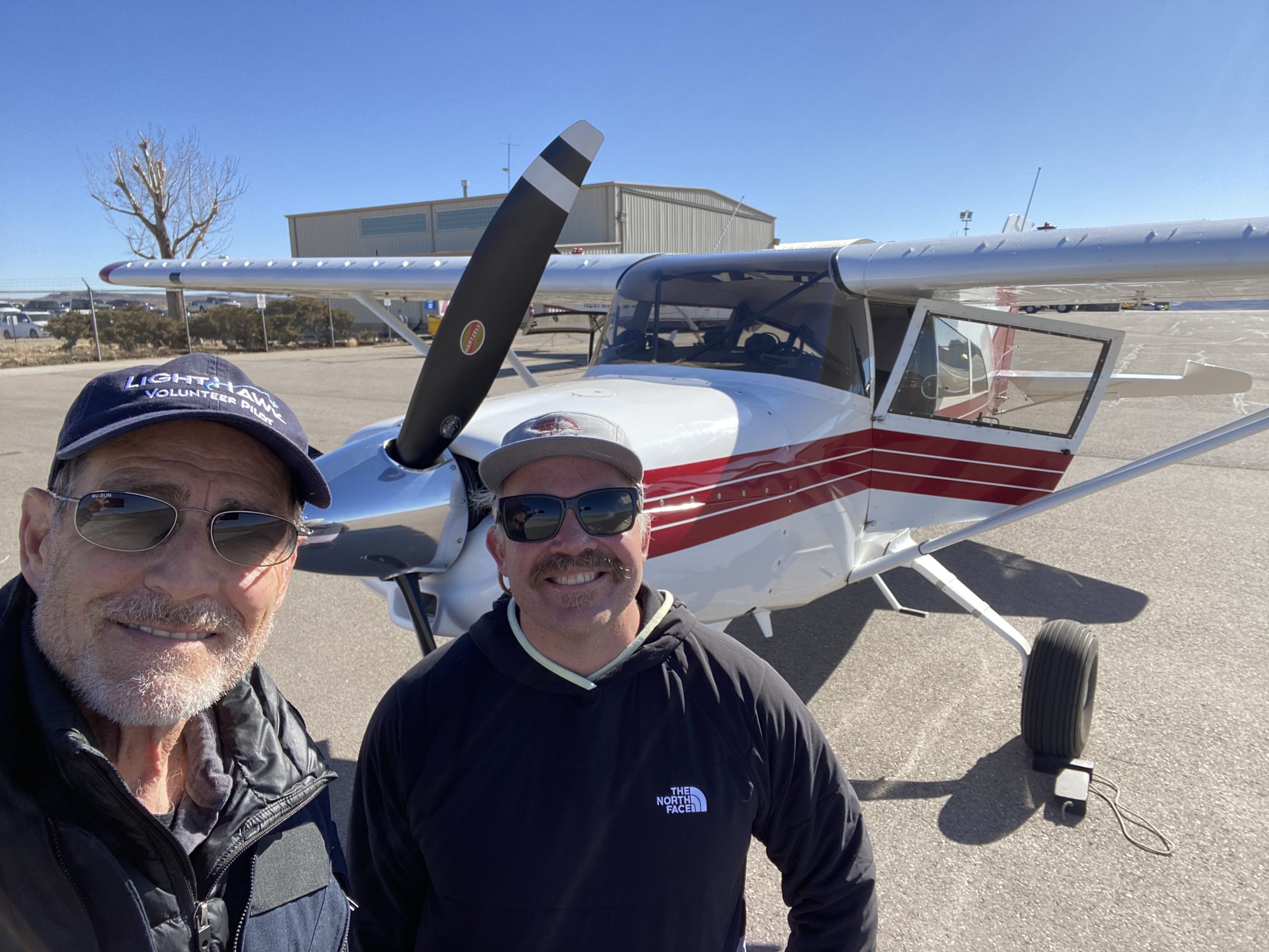 Two men standing in front of a small, high-wing aircraft.